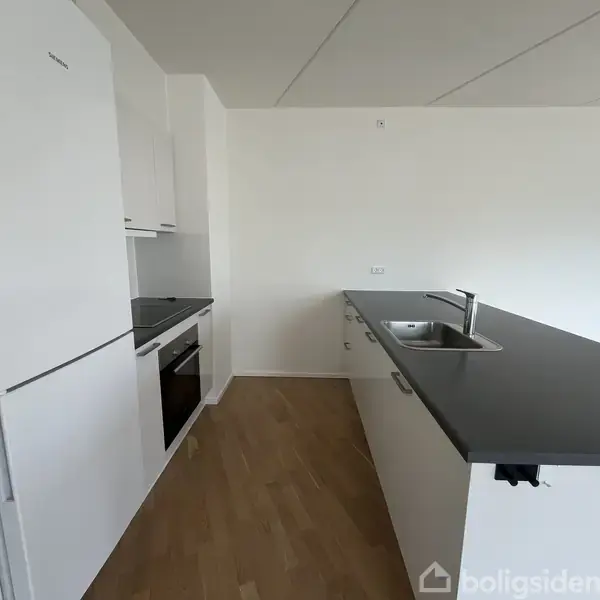 Køkken med hvide skabe og sort bordplade rummer indbyggede apparater som ovn og køleskab. Der er en vask i øen. Trægulv omgiver området. Vægge er hvide.