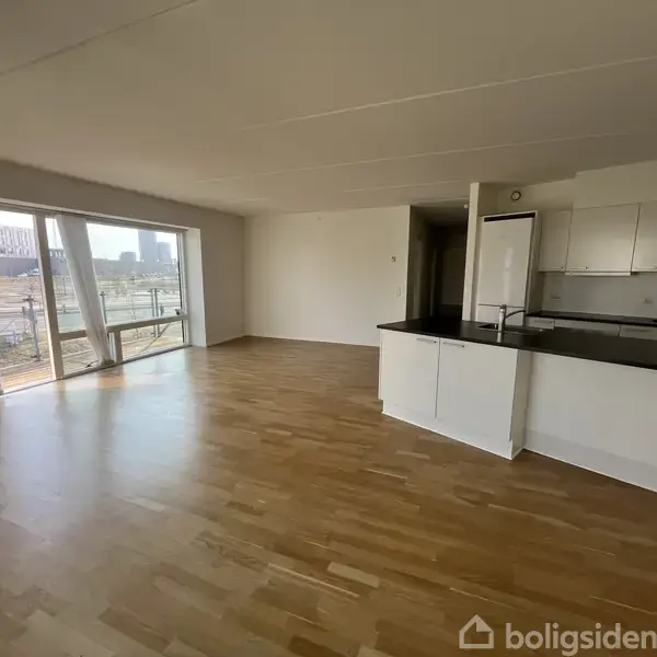 Lejlighedsstue med parketgulv. Køkken med hvidt interiør på højre side. Store gulv-til-loft vinduer til venstre giver udsigt over en by.