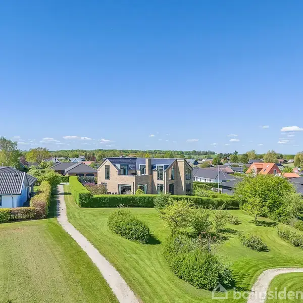 Et moderne hus står i en grøn, velplejet have. En sti fører op til huset, omgivet af andre huse i et villakvarter under en klar blå himmel.