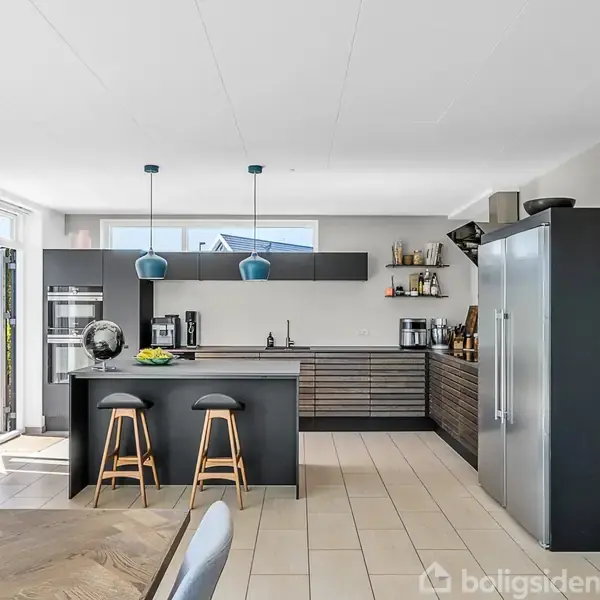 Køkken med mørke skabe og skuffer arrangeret langs væggene, barstole foran en central ø, lamper hænger over, store vinduer oplyser rummet, moderne kunst hænger på væggen.
