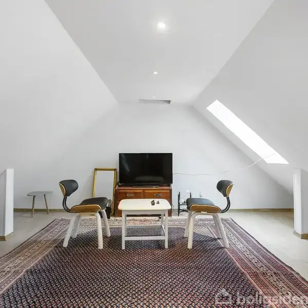 Et fjernsyn står på en kommode med stole foran, beliggende i et loftsrum med hvide vægge og skråt tag. Et stort tæppe dækker gulvet.