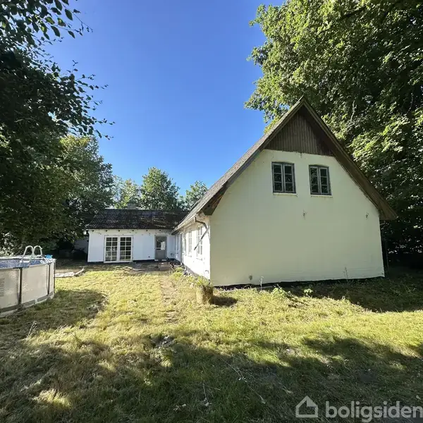 Et hvidt hus står stille i en solrig have omkranset af træer; en rund, hævet pool ses til venstre.