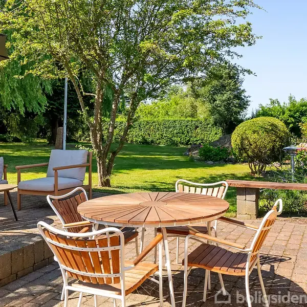 Rundt havebord med træstole står på en terrasse med fliser. En sofa og parasol ses til venstre. Haven er grøn med træer og buske i baggrunden.