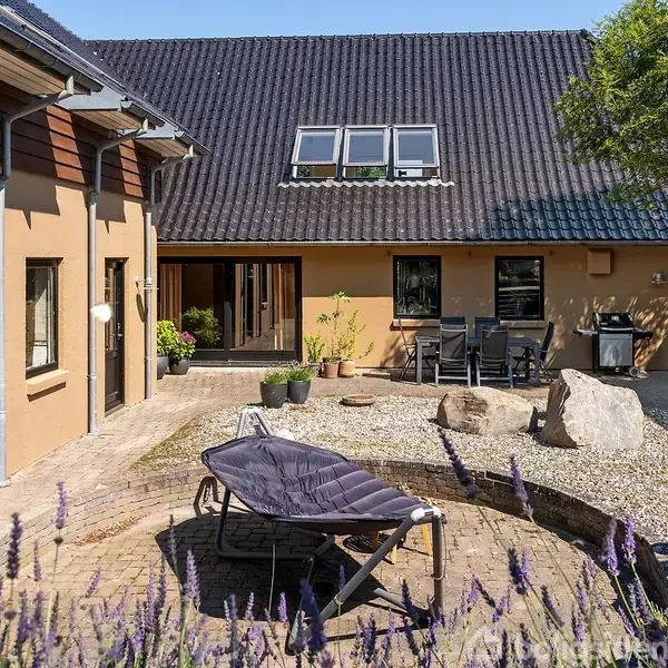 Solstol står i en gårdhave omgivet af lavendel og sten. Til højre er der havemøbler og grill ved en beplantet væg. Huset har ovenlysvinduer og mørkt tag.