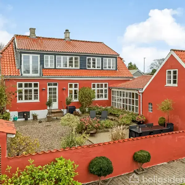 Et rødt murstenshus står i en baggård med en gårdhave fyldt med planter og udendørs møbler. Huset har hvide vinduer og et orangetag, omkranset af en mur med buegang.