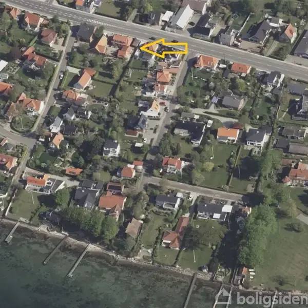 Et luftfoto viser et beboelseskvarter nær en kystlinje. Huse med røde tage ligger langs veje, og en gul pil peger mod et bestemt hus. Vandet ses nederst i billedet.