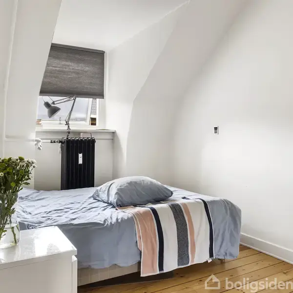Seng er redt med blåt sengetøj i et lille hvidmalet rum, placeret ved skrånende vægge. Et natbord med blomster står ved siden af, og et vindue med rullegardin er bagved.