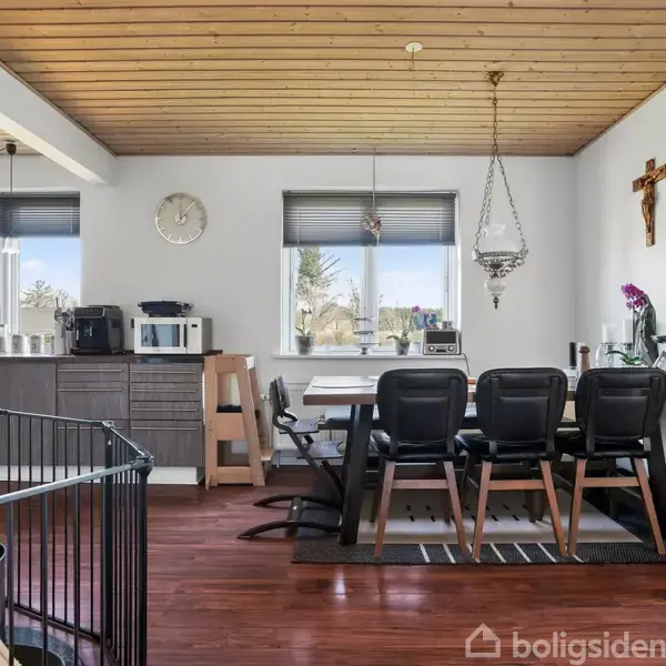 Et køkken-alrum med spisebord og seks stole. På væggen hænger et krucifiks og en lampe, og der er udsigt gennem vinduer. Klokken på væggen viser tiden, og planter står i hjørnet.