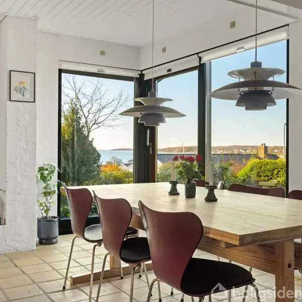 Et spisebord med fire stole står i et lyst rum med flisegulv og store vinduer. En pejs er til venstre, og der er udsigt til en have og vand i baggrunden.