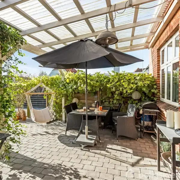 Murstenshus med rødt tag står i en grøn have. En flisebelagt terrasse med havemøbler til venstre. I baggrunden er nabohuse synlige under en blå himmel.