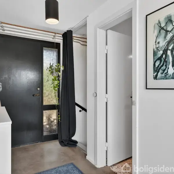Dør er sort, lukket og omgivet af lys gennem glasrude. Skænk til venstre med vase og blomster. Hvid væg med kunst hænger til højre. Gardiner dækker vinduerne.