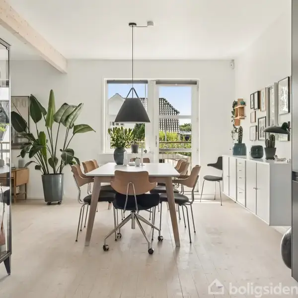 En spisestue med et rektangulært bord og seks stole. En sort pendellampe hænger over bordet. Store planter står i rummet, og lyse vægge er dekoreret med billeder.