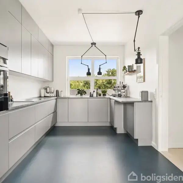 Moderne køkken med grå skabe, indbygget ovn, og sort gulv. Store vinduer slipper naturligt lys ind. To sorte, geometriske lamper hænger i loftet. Planter og køkkenredskaber på bordplade.