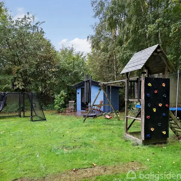 Legetårn med klatrevæg står i en grøn have. Bagved ses en blå skur og en trampolin. Omgivet af buske og træer, skaber det en hyggelig udendørs atmosfære.