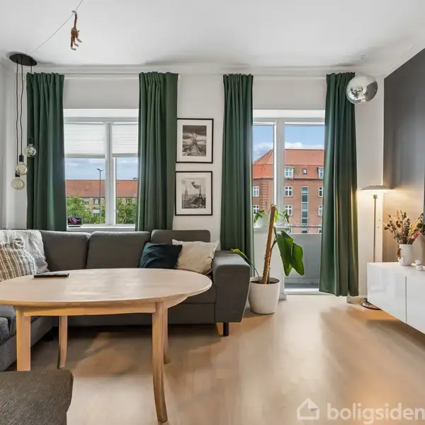 Stue med grå hjørnesofa og rundt træbord, placeret ved store vinduer med grønne gardiner. Væggene har kunsttryk, og der står en plante og lamper omkring rummet.