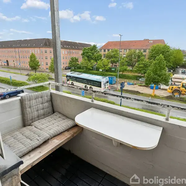 Balkon med en pude-klædt bænk og klapbord. Udsigt over en vej med biler, en bus ved et stoppested og en gravko nær en bygning omgivet af træer og boliger.