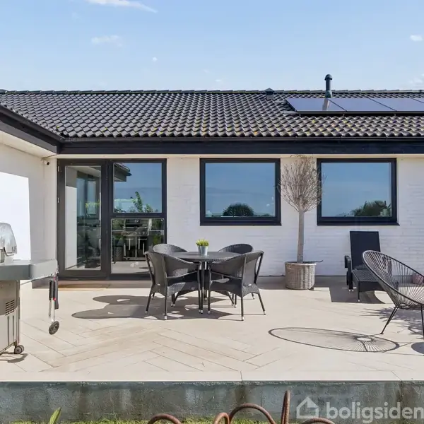En terrasse med et rundt bord og fire stole står foran et hvidt hus med sorte tagsten og solpaneler. En grill er placeret til venstre på terrassen.