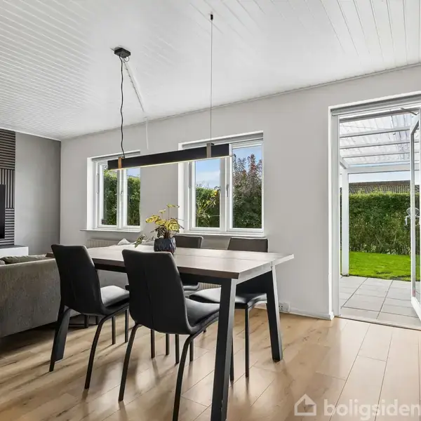 En spisestue med et bord og fire stole, hvor en lampe hænger over bordet. Til venstre er sofaen foran et TV. Døren åbner ud til en terrasse og grøn have.