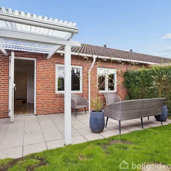 Terrasse med havemøbler står på fliser foran et rødt murstenshus. Dør er åben til huset, omgivet af grønne buske og græsplæne i baggrunden.