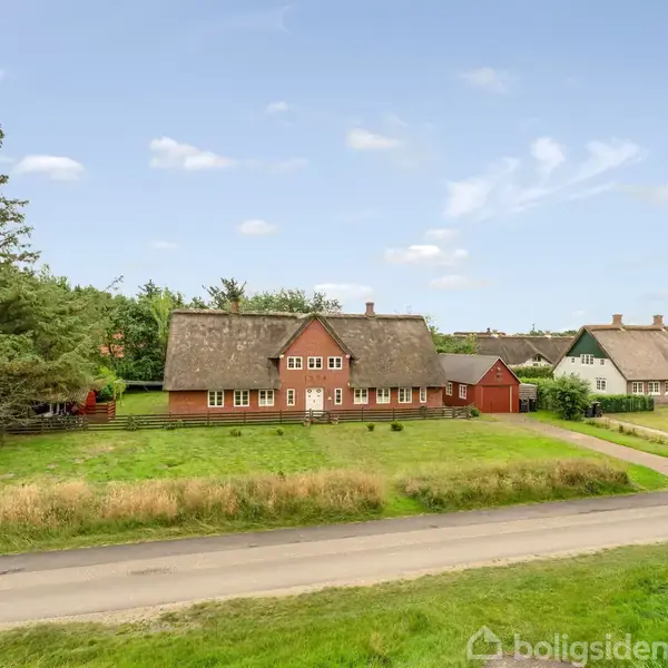 Rødt bindingsværkshus med stråtag står stille på en grøn grund. Omkringliggende træer og andre huse danner en landlig landskabsbaggrund. En vej løber foran huset.
