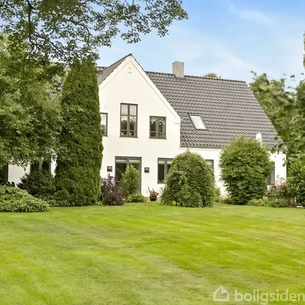 Et hvidt hus med sort tag står i en velplejet have med grøn græsplæne, omkranset af høje træer og buske. Ingen tekst er synlig.