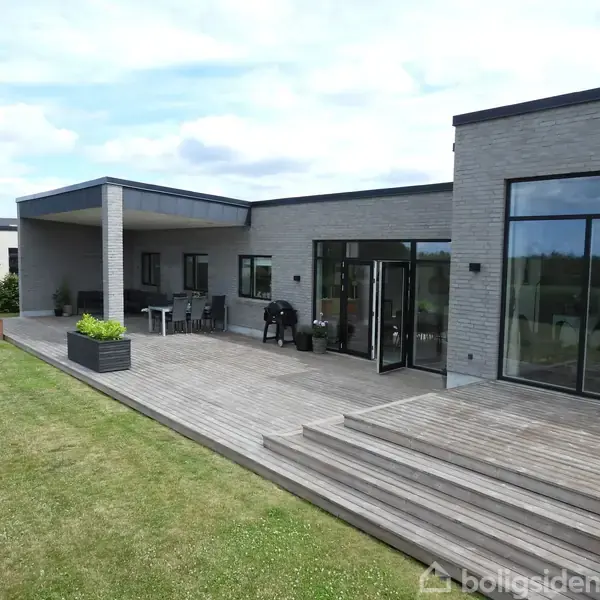 Et moderne hus med fladt tag står stille omgivet af en velplejet græsplæne og en asfalteret vej. I baggrunden ses åbne marker og et skovklædt landskab.