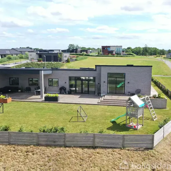En moderne villa med fladt tag står på en velplejet græsplæne. En legeplads med rutsjebane er i haven, omgivet af et hegn. Baggrunden viser et beboelsesområde.