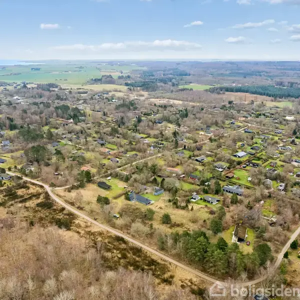 Landsbyområde med huse spredt blandt træer, omgivet af åbne marker og skove under en klar blå himmel med lette skyer.