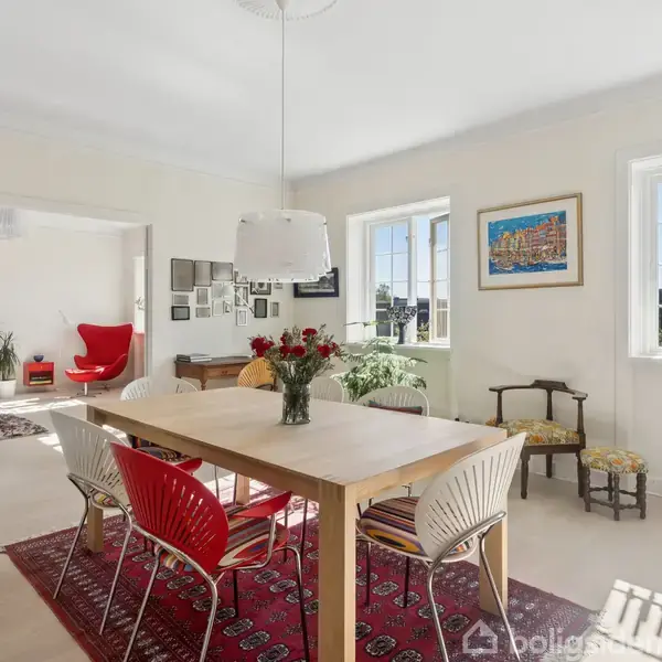Et spisebord med stole står på et rødt tæppe i en lys stue. Bordet er dekoreret med en vase blomster. Væggene har kunstværker, og store vinduer giver naturligt lys.