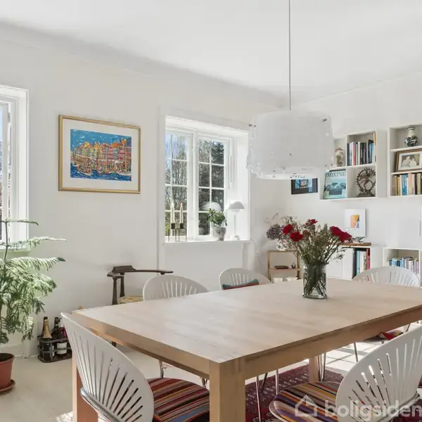 En spisebord med vase og blomster står i et lyst rum. Hvide stole omringer bordet. Væggene har indbyggede bogreoler, kunstværker, og der er to store vinduer.