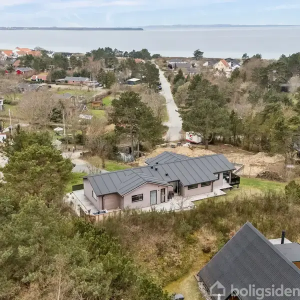 Et moderne hus med gråt tag beliggende i et naturskønt område med træer og buske. I baggrunden ses flere huse og en vej, der fører mod en fjord.