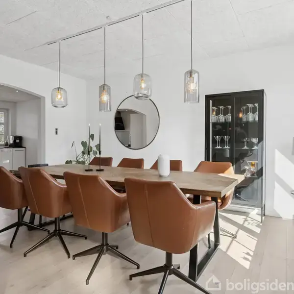 Et spisebord med brune læderstole omkring står i et lyst spisestueområde. Over bordet hænger tre moderne lamper; i baggrunden ses en skænk med glas og en rund spejl.