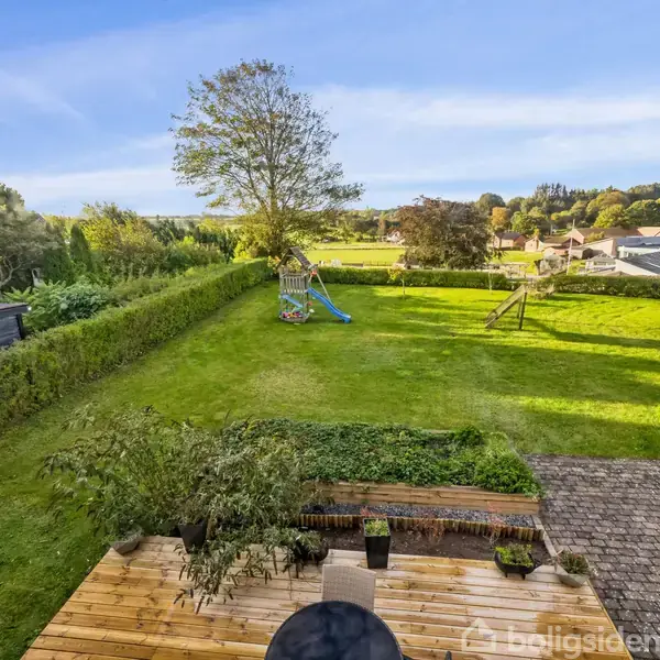Terrasse omgivet af grøn have med gyngestativ og rutsjebane. Hæk danner grænse til åbne marker og videre til en boligområde i baggrunden under en blå himmel.