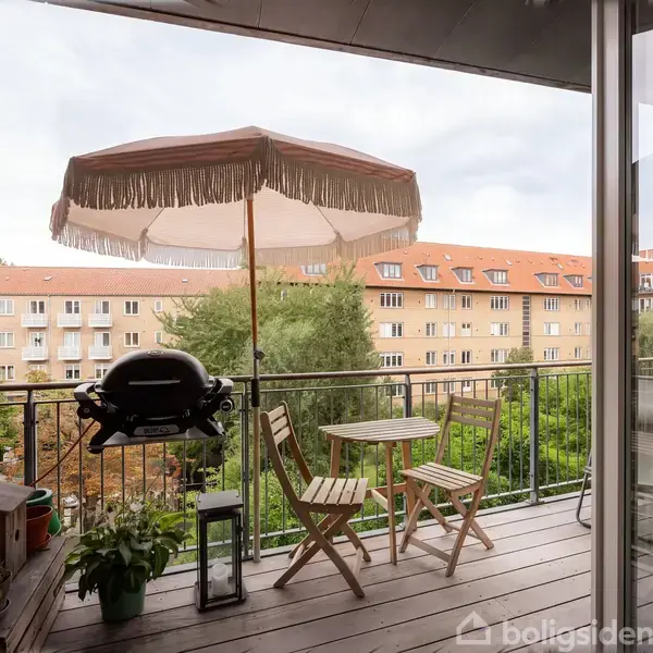 Balkon med træmøbler og parasol. Grill står på rækværket. Omgivet af murstensbygning og udsigt til gule lejlighedskomplekser. Planter tilføjer grønne elementer.