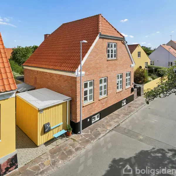 Et rødt murstenshus med rødt tegltag står langs en stille vej med fortov. Huset har flere hvide vinduer og en tilstødende gul bygning. Solskin og blå himmel ses over området.