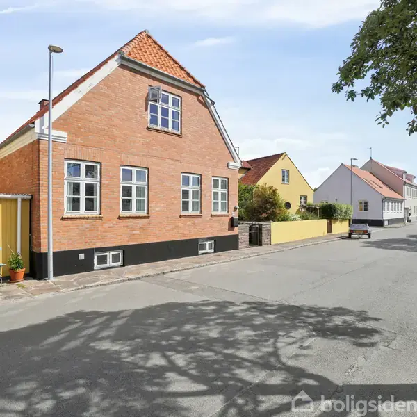 En rød murstensbygning med hvide vinduer står langs en stille, træbevokset vej i et boligområde. Andre farverige huse ses i baggrunden under en klar blå himmel.