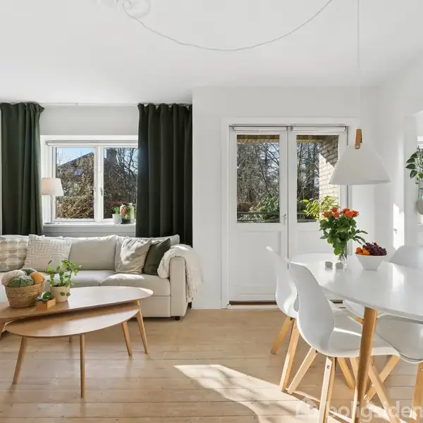 Sofaen står ved siden af et lyst og åbent vindue, mens et rundt sofabord bærer planter. En hvid spisebordssæt med blomster indrammes af hvide vægge og trægulve.