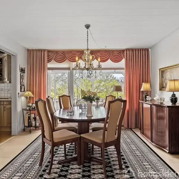 Et spisebord med seks stole står på et tæppe i en elegant spisestue. En lysekrone hænger over bordet, omgivet af skabe og lamper. Store vinduer viser en have.