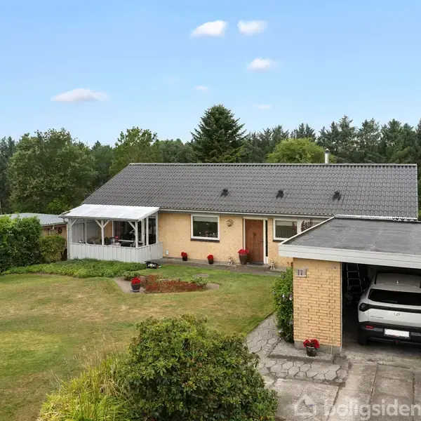 En etplanshus med grå tagsten står for enden af en anlagt have og en carport med en bil. Omkring huset er der græsplæner og buske.