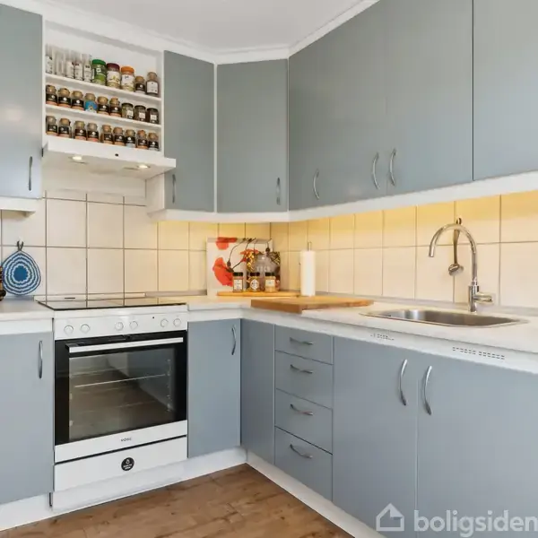 Et køkken med blågrå skabe omgiver en hvid ovn og komfur. Over ovnen er en hylde med krydderiglas. Til højre er en vask med hane. Køkkenredskaber pryder bordpladen.