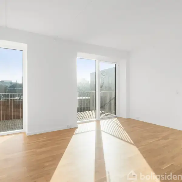 Lyst rum med trægulv og store vinduer, åbner ud mod en balkon. Sollys strømmer ind, hvilket skaber klare skygger. En del af et moderne, minimalistisk indretningsdesign.