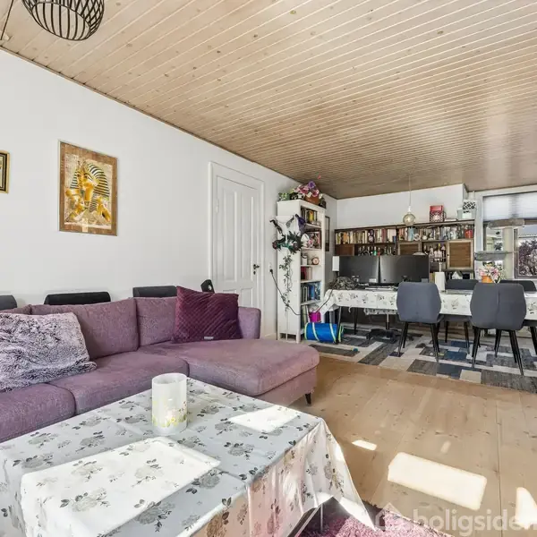 Lilla sofa med puder står i en stue ved siden af et rektangulært sofabord med dug. Stuen har trægulv og -loft samt spiseområde med dækket bord og stole i baggrunden. Vægge er pyntet med billeder.