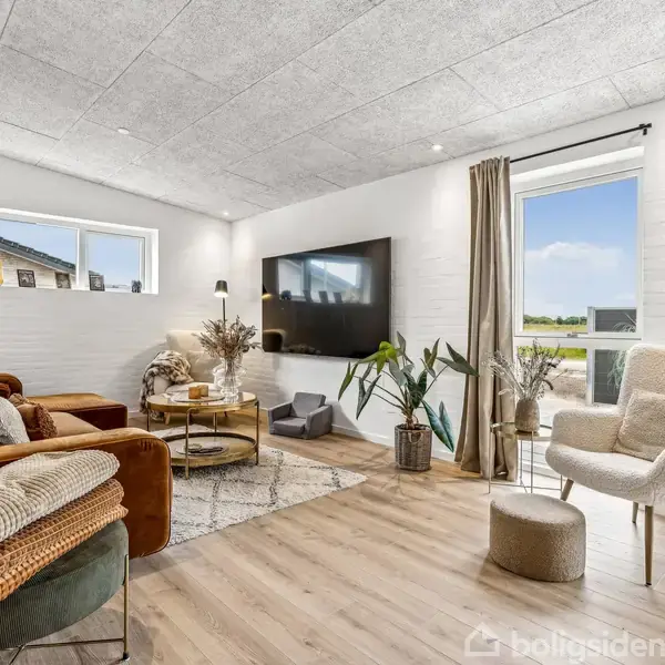 Stue med brun sofa og lænestol, dekoreret med planter og tæpper. Fladskærms-tv hænger på væggen. Store vinduer med lysindfald giver kig til natur udenfor.