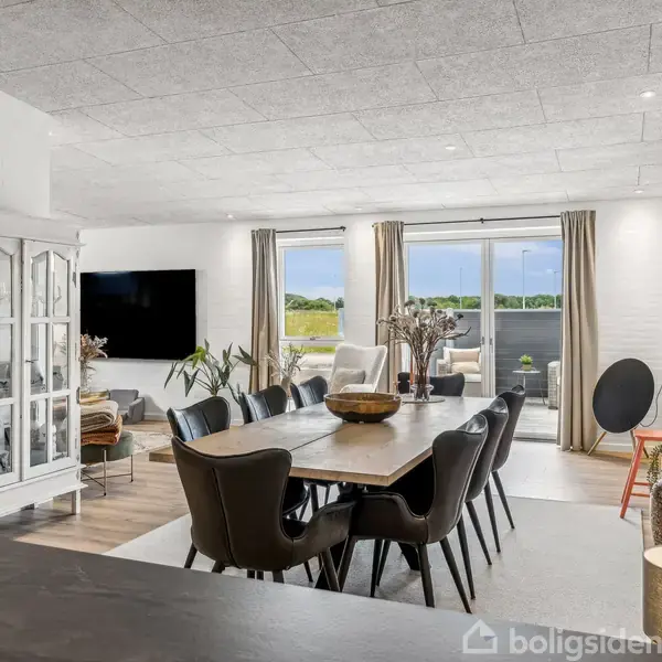 En spisestue med stort bord og polstrede stole placeret i en lys og moderne stue med store vinduer, der åbner ud til en terrasse. En hvid vitrineskab står ved siden af, og en stor plante ses i rummet.