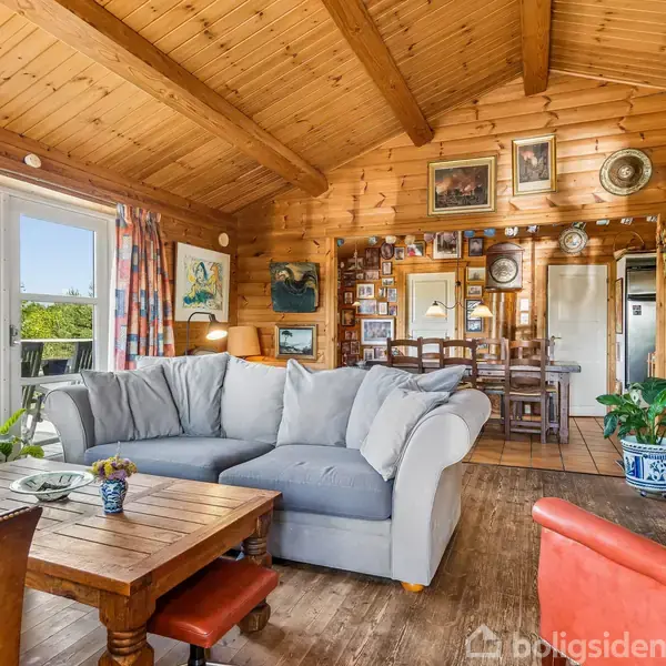 Grå sofa står i en stue med trævægge og -loft. Stuen er dekoreret med billeder og vægure. Store vinduer med udsigt til grønne omgivelser. Borde og stole fuldender rummet.