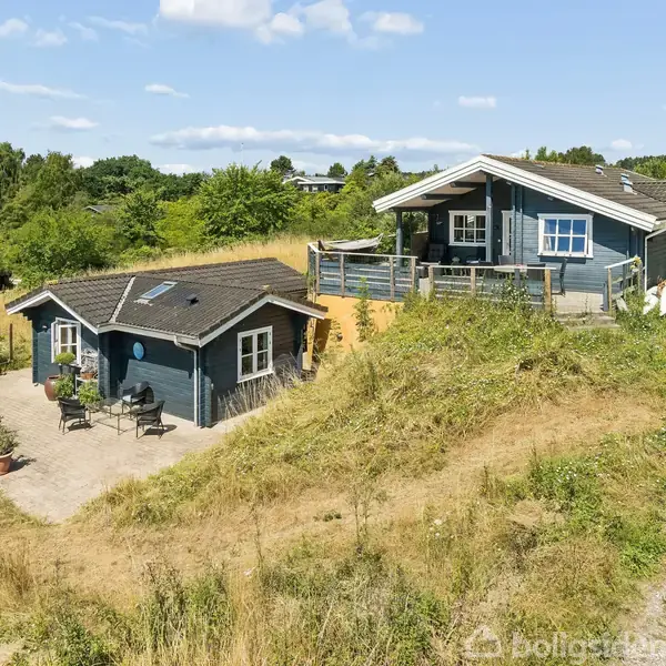 To blå sommerhuse med terrasser står i et bakket landskab med stier og grønt buskads. En gårdhave foran det lille hus har havemøbler.