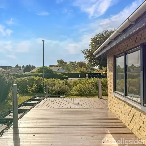 En træterrasse ved siden af et hus med gule mursten, omgivet af grønne buske. Terrassen er tom, og der er udsigt til en rolig, beboet omgivelse under en blå himmel med skyer.