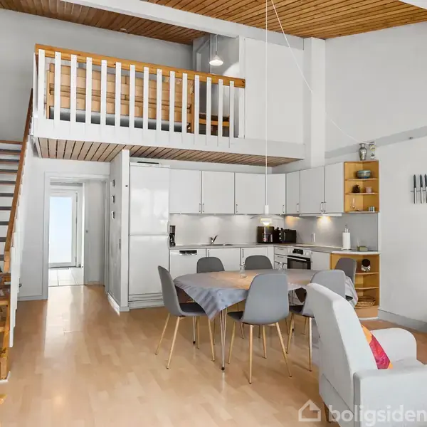 Trætrappe fører op til hems i lys, åben stue. Hvidt køkken med spisebord og grå stole. Sofa med puder placeret til højre. Loftsrummet har trægulv og hvidmalede vægge.