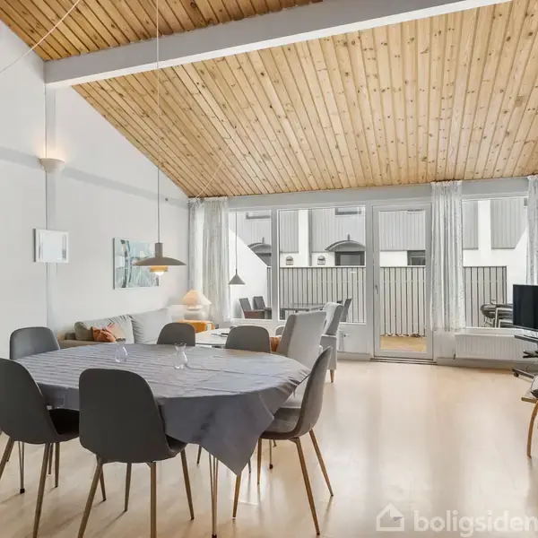 En spisestue med et rundt bord dækket med en dug og omgivet af seks stole. I baggrunden er der en sofa og fladskærms-tv. Træpaneler pryder loftet.