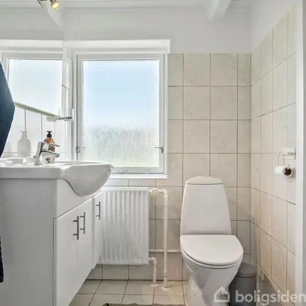 Et lyst badeværelse med toilet og vask placeret ved siden af hinanden, omgivet af lyse fliser. Et stort vindue lader naturligt lys komme ind. En radiator står under vinduet.